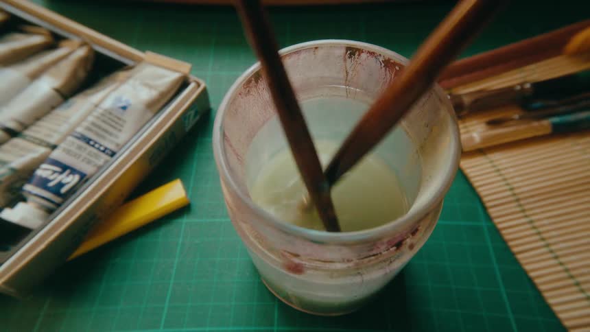 An artist cleans a paintbrush in a cup of water on a workbench with art supplies