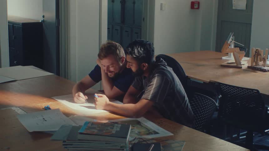 Two creative male partners collaborate on a project, sketching ideas together on a large paper in a sunlit studio