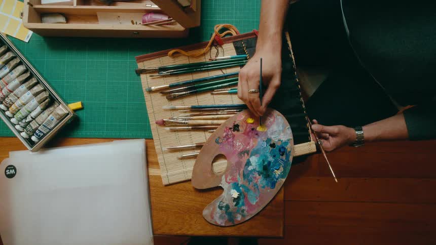 An overhead shot shows an artist mixing vibrant yellow oil paint on a wooden palette in a cozy studio setting