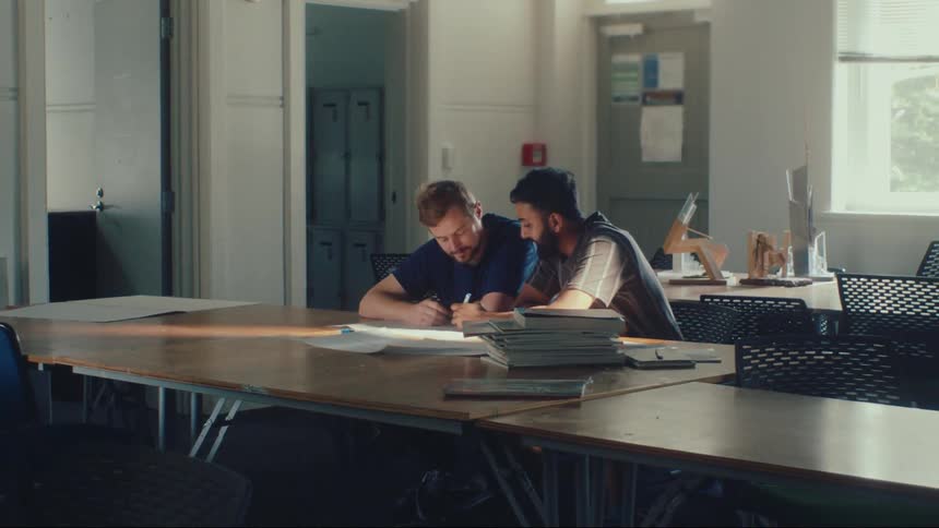 Two friends and colleagues work together, happily drawing on a big piece of paper at a large table in a bright classroom