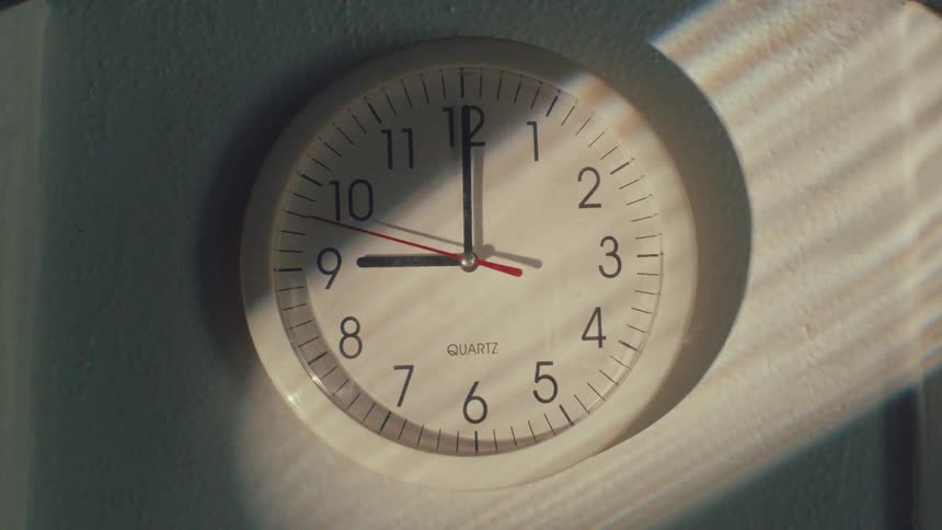 A white analog wall clock at nine o'clock with sunlight and shadows moving across its face