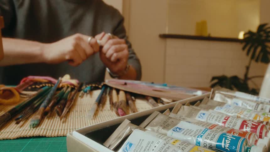 An artist carefully selects a tube of yellow oil paint from a box and squeezes it onto a wooden palette in slow motion