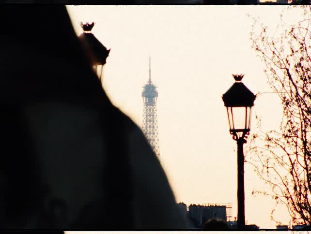 A vintage film shot of the Eiffel Tower framed between two silhouetted street lamps as people walk by at sunset in Paris, France
