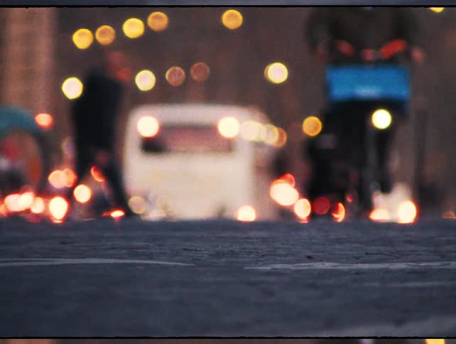 Out-of-focus, low-angle shot captures the bustling city life at night as cars, bicycles, and pedestrians move past in a blur of motion and light