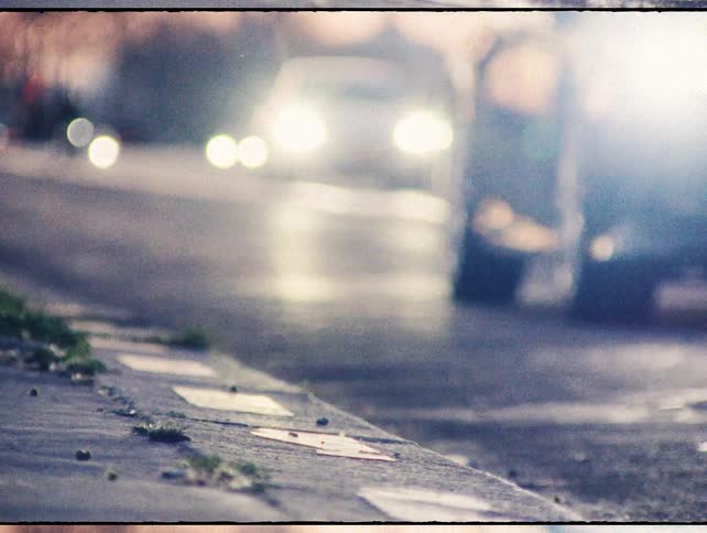 A gritty, low-angle shot from a street curb shows blurry car headlights passing by during dusk