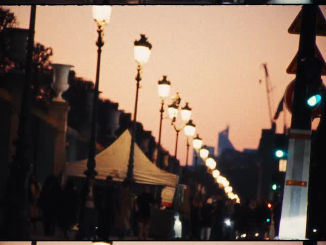 Vintage film captures a bustling Parisian street scene at dusk, with a row of glowing street lamps under a warm, pink sky