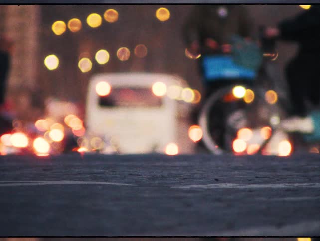 An artistic low-angle, out-of-focus shot captures cyclists and pedestrians moving along a city street against a backdrop of glowing traffic lights at dusk