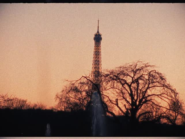 A grainy vintage film shot shows the Eiffel Tower in Paris silhouetted against a warm sunset sky with a fountain in the foreground
