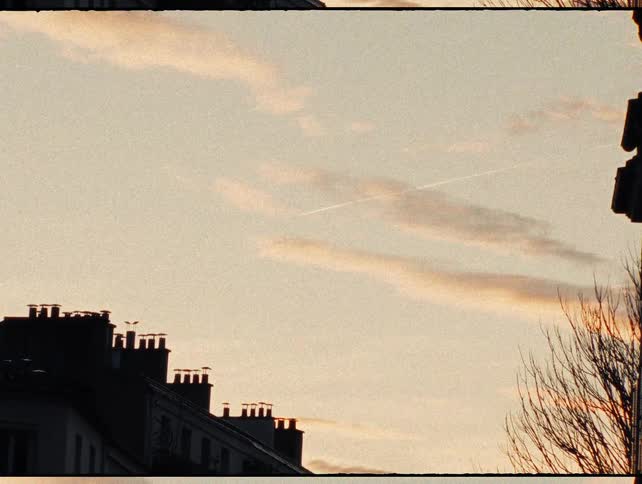 A grainy, vintage-style shot zooms out from a jet trail in the sky, revealing a beautiful sunset between two silhouetted city buildings