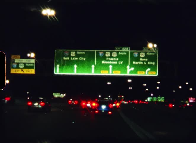 Point-of-view shot driving on a busy Las Vegas freeway at night, passing under a glowing green road sign with a vintage film aesthetic