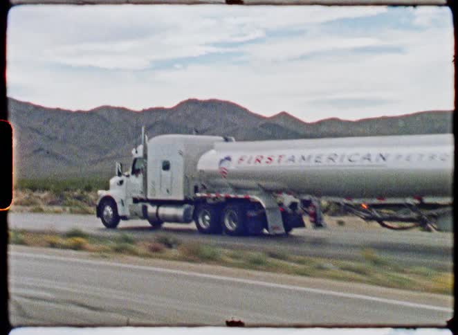 A vintage, grainy film clip captures a First American Petroleum tanker truck driving along a desert highway with mountains in the distance