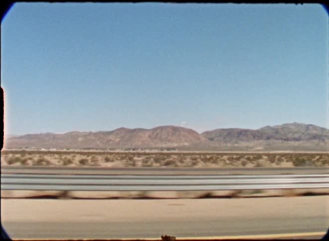 A view from a moving car captures traffic passing on a desert highway with mountains in the distance, shot on vintage film