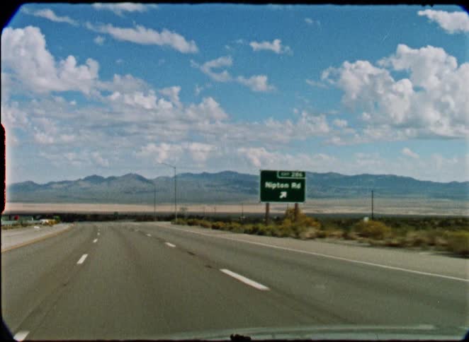 A vintage, point-of-view shot from a car driving on a desert highway towards distant mountains under a cloudy blue sky