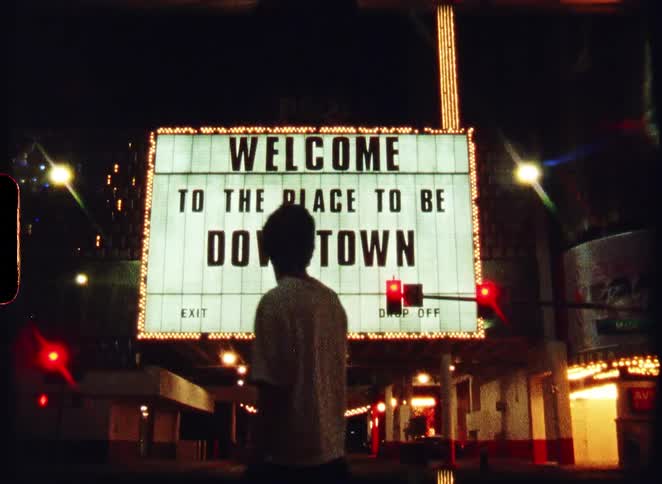 Retro-style 8mm film shows a person admiring a glowing 'Welcome To The Place To Be Downtown' sign on a city street at night