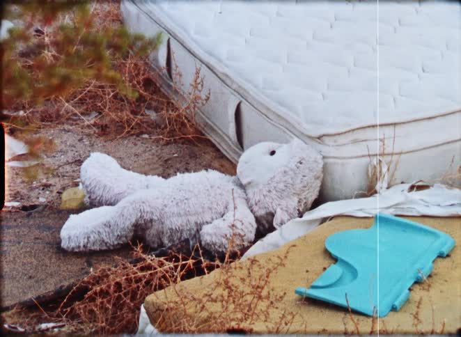A lonely, discarded white stuffed animal lies on the ground next to an abandoned mattress in a desolate area, captured on vintage film