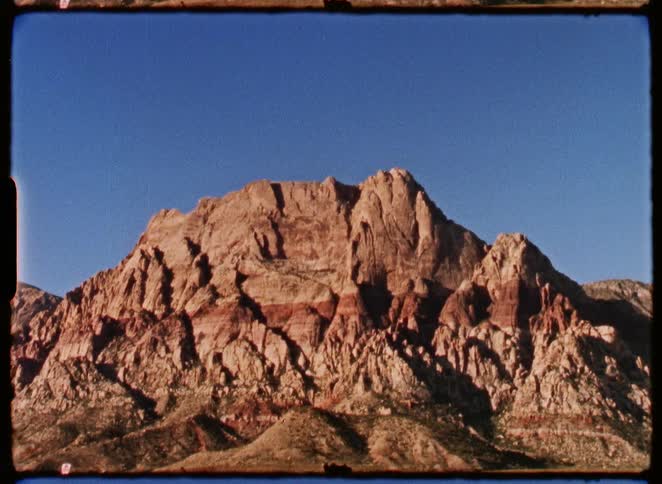 A static shot of a rugged, sunlit mountain formation against a deep blue sky, captured on grainy vintage film