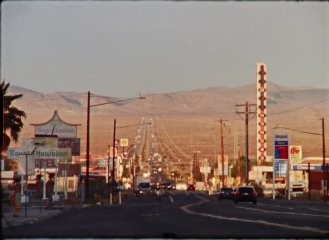 A vintage film view of the long, straight road through Baker, California, featuring the World's Tallest Thermometer at golden hour