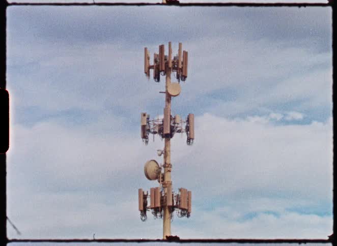 A stationary shot of a cellular tower against a cloudy blue sky, captured on grainy vintage film