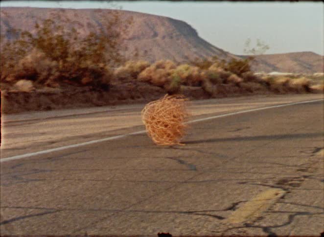 Vintage footage shows a lone tumbleweed rolling across a cracked and empty desert road with mountains in the background