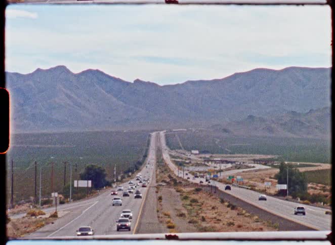 A long view of a desert highway with cars traveling towards distant mountains, captured on vintage film