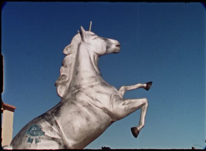 A quirky, silver unicorn statue with a Pabst Blue Ribbon beer logo on its flank, shot on vintage 8mm film against a clear blue sky