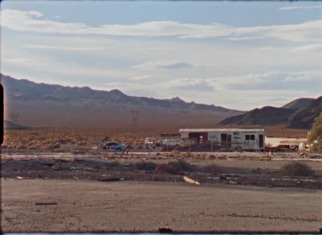 Vintage Super 8 film captures a desolate view of an abandoned trailer and wrecked cars in a remote desert landscape