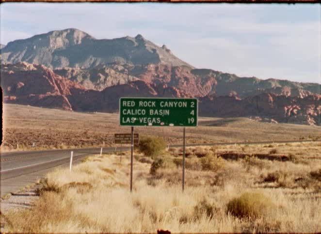 A retro film captures a roadside sign for Red Rock Canyon and Las Vegas amidst the vast Nevada desert landscape