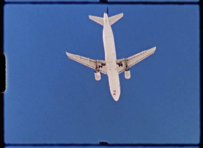 A vintage, low-angle film shot captures a passenger jet flying overhead against a clear blue sky