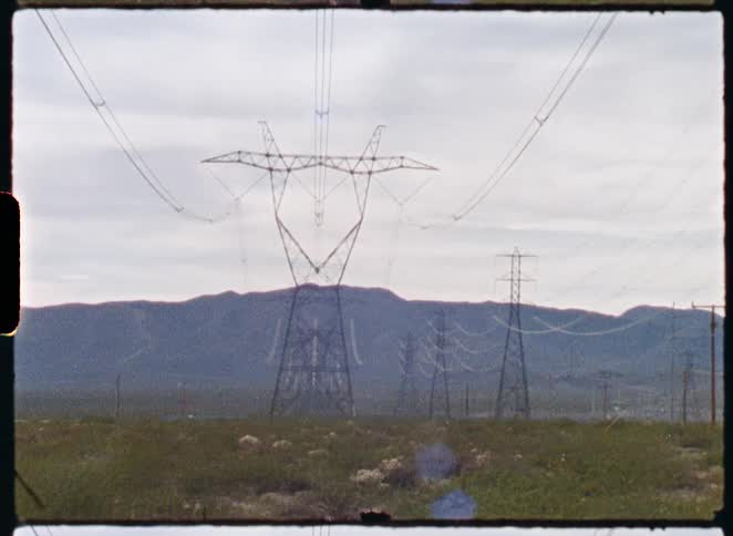 A vintage film shot captures electrical transmission towers stretching across a vast, arid landscape with distant mountains under an overcast sky