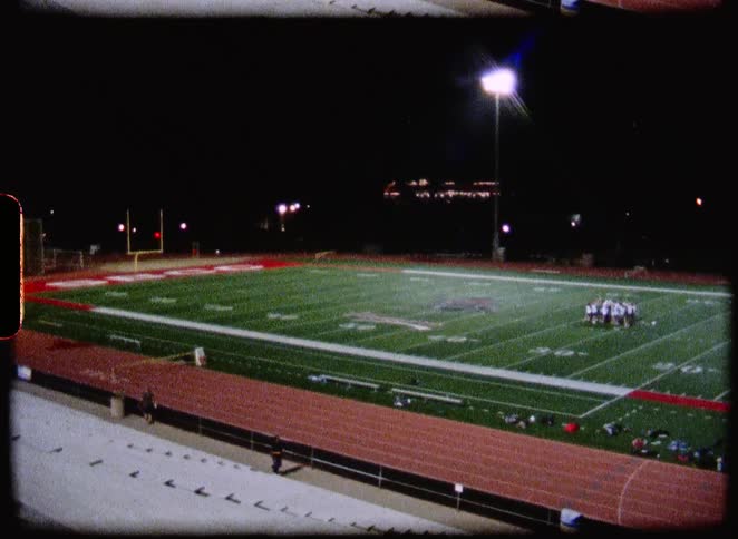 A vintage wide shot of a football team huddled on a brightly lit field at night