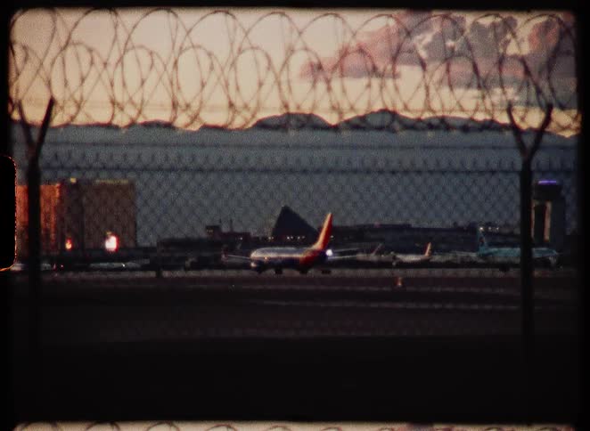 Vintage 8mm footage captures a plane taxiing on the runway at the Las Vegas airport at dusk, viewed through a barbed wire fence