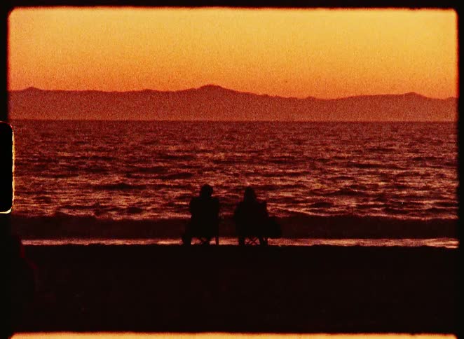 A silhouetted couple enjoys a romantic and peaceful sunset over the ocean from their beach chairs in this vintage film footage