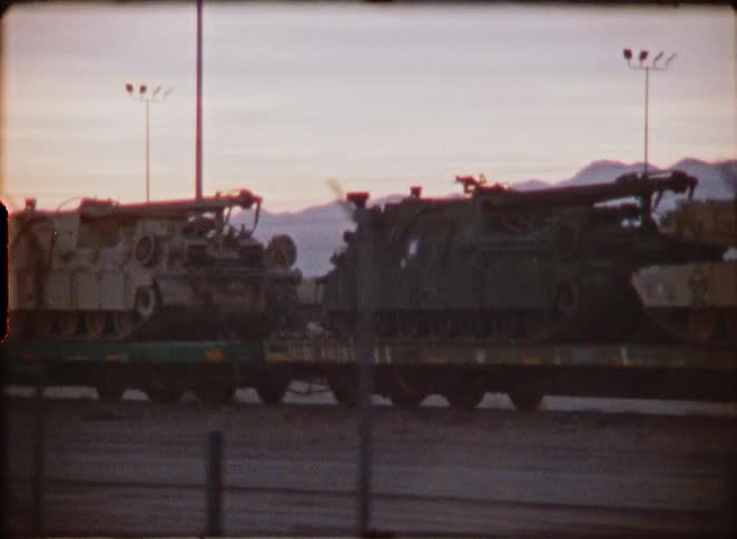 Vintage 8mm footage shows a train transporting military tanks and armored vehicles through the desert at dusk