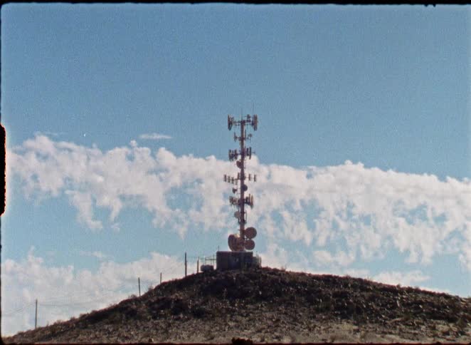 A slow zoom on a remote communications tower atop a barren desert hill against a blue sky, shot on 8mm film