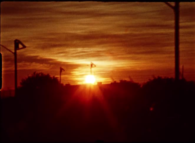 A vintage super 8mm film captures a warm, glowing sunset over a silhouetted industrial landscape