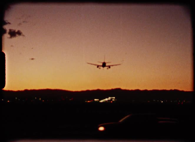 Vintage 8mm footage captures an airplane silhouetted against a colorful sunset sky as it descends for landing