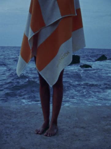 A person wrapped in a striped orange and white towel stands barefoot on a concrete pier overlooking a choppy blue sea
