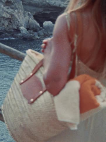 A woman with a woven beach bag and rolled-up towel stands on a cliff overlooking the ocean