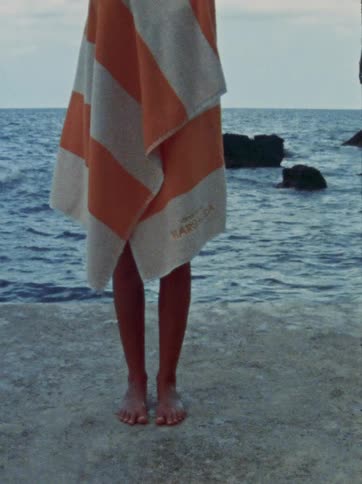 A person wrapped in a striped orange and white towel stands barefoot on a concrete pier overlooking the calm ocean