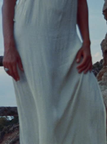 A vintage-style shot of a woman in a breezy white dress standing on a rocky cliff as the wind blows