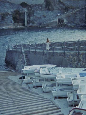 Vintage 8mm footage captures a woman in a towel walking along a stone pier next to docked boats at dusk