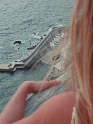A blonde woman looks down from a high vantage point at a scenic coastal harbor with boats docked on a stone ramp