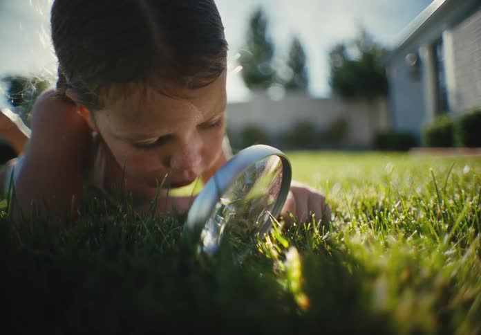 A curious young girl lies on her stomach in the lush green grass, intently examining it with a magnifying glass on a sunny day