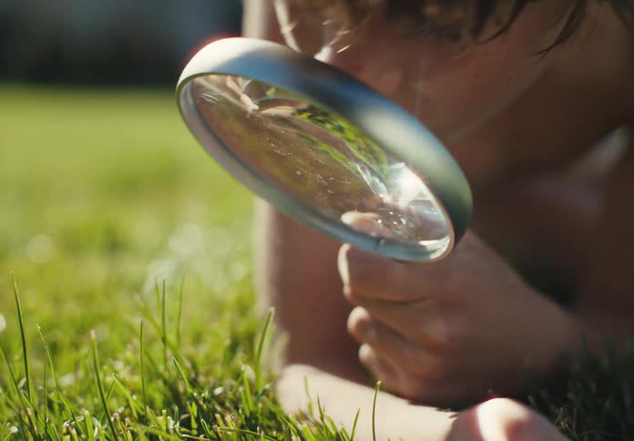 A curious boy with wet hair lies in the grass and looks through a magnifying glass on a sunny day