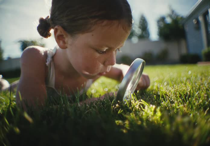 A curious young girl lies in the bright green grass, intently exploring nature with a magnifying glass on a sunny day