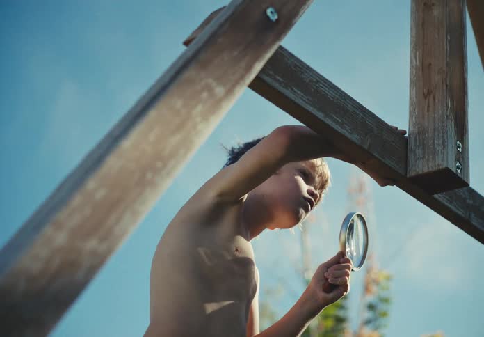 Low angle shot of a curious young boy intently examining a wooden structure with a magnifying glass under a clear blue sky