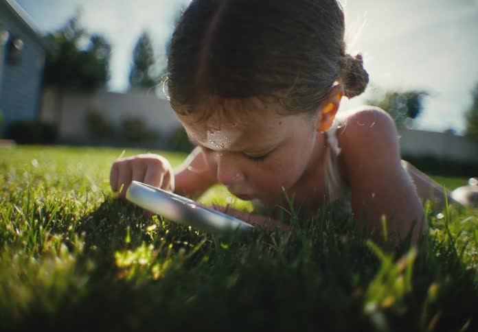 A curious young girl lies on a bright green lawn, intently exploring the grass with a magnifying glass on a sunny day