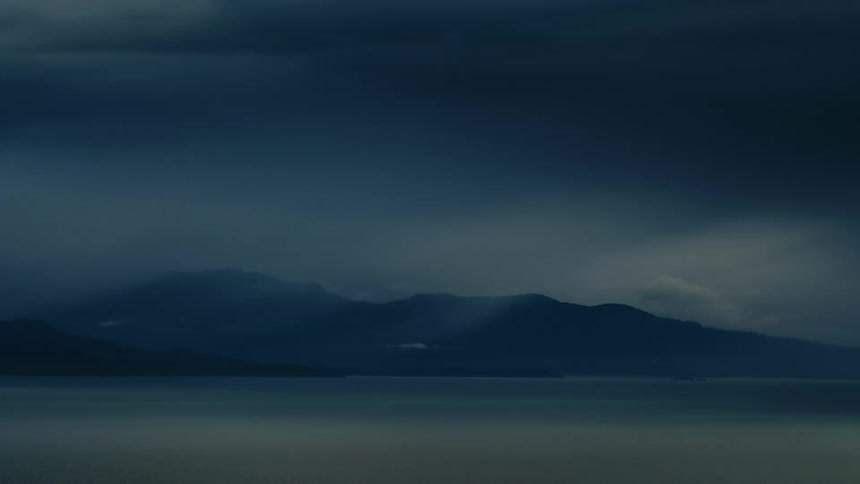 Dark storm clouds and fog roll over a mountain range across a calm body of water
