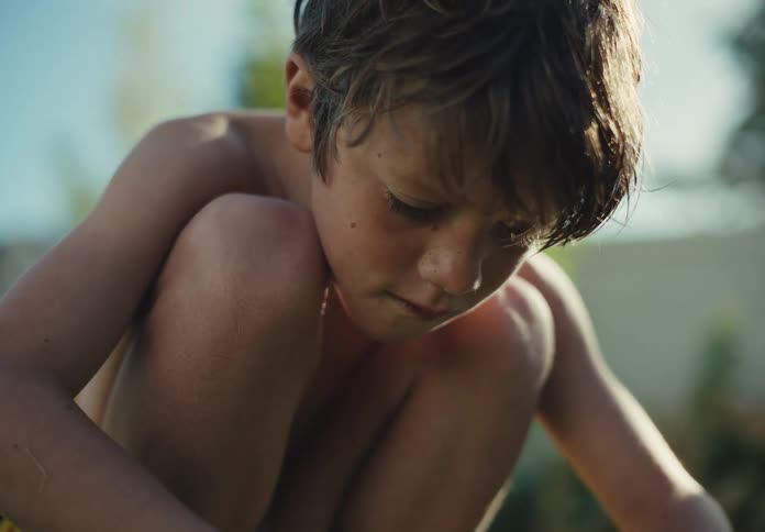 Young boy crouches outdoors in warm sunlight with a focused and contemplative expression