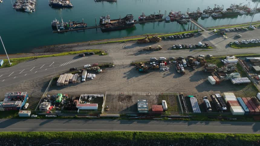 A static aerial view shows an industrial port yard with equipment and containers alongside a harbor full of docked boats